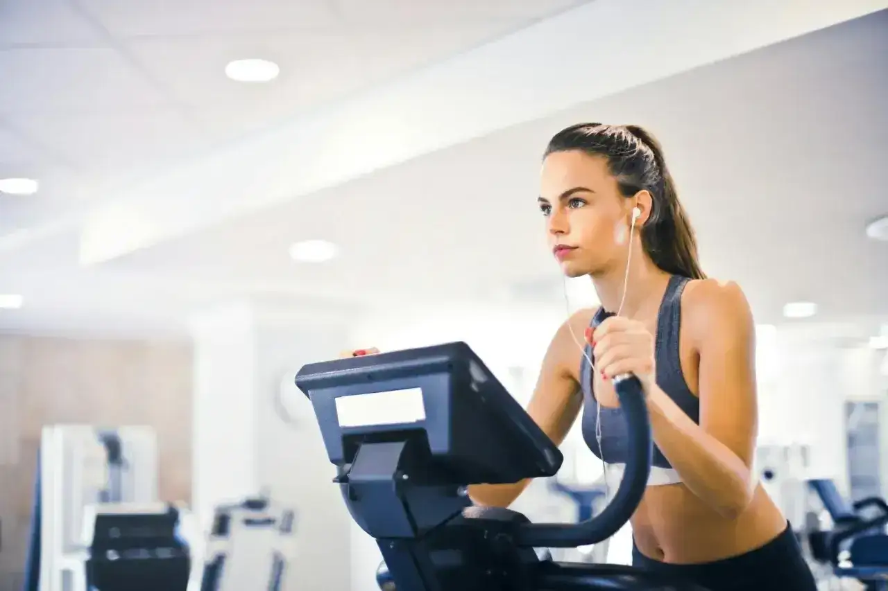 Kobieta w słuchawkach wykonuje cardio po treningu siłowym na bieżni eliptycznej w siłowni.