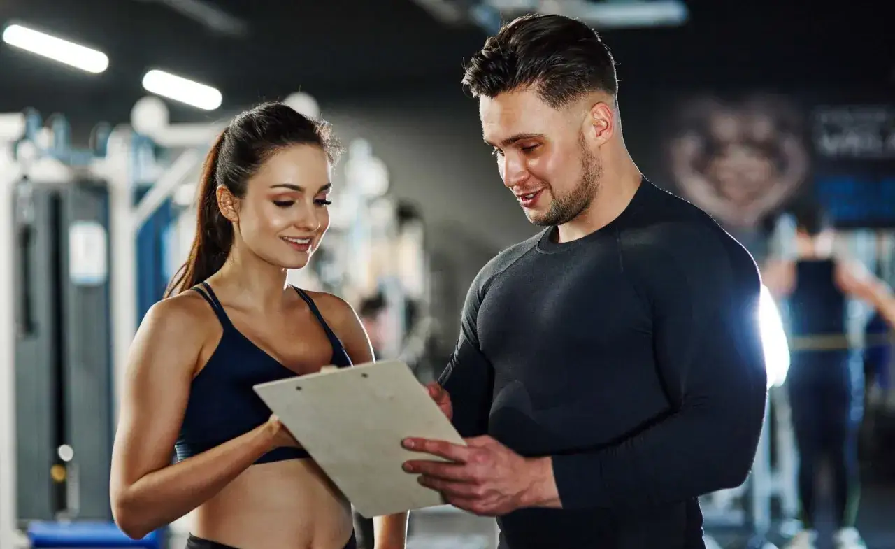Ein Mann und eine Frau besprechen Trainingspl&auml;ne im Fitnessstudio. Sie sind beide sportlich gekleidet. Die Frau h&auml;lt eine Mappe. Der Mann scheint ihr etwas zu erkl&auml;ren. Die Szene k&ouml;nnte die Ausbildung eines Fitnesskaufmanns und das damit verbundene Geh...