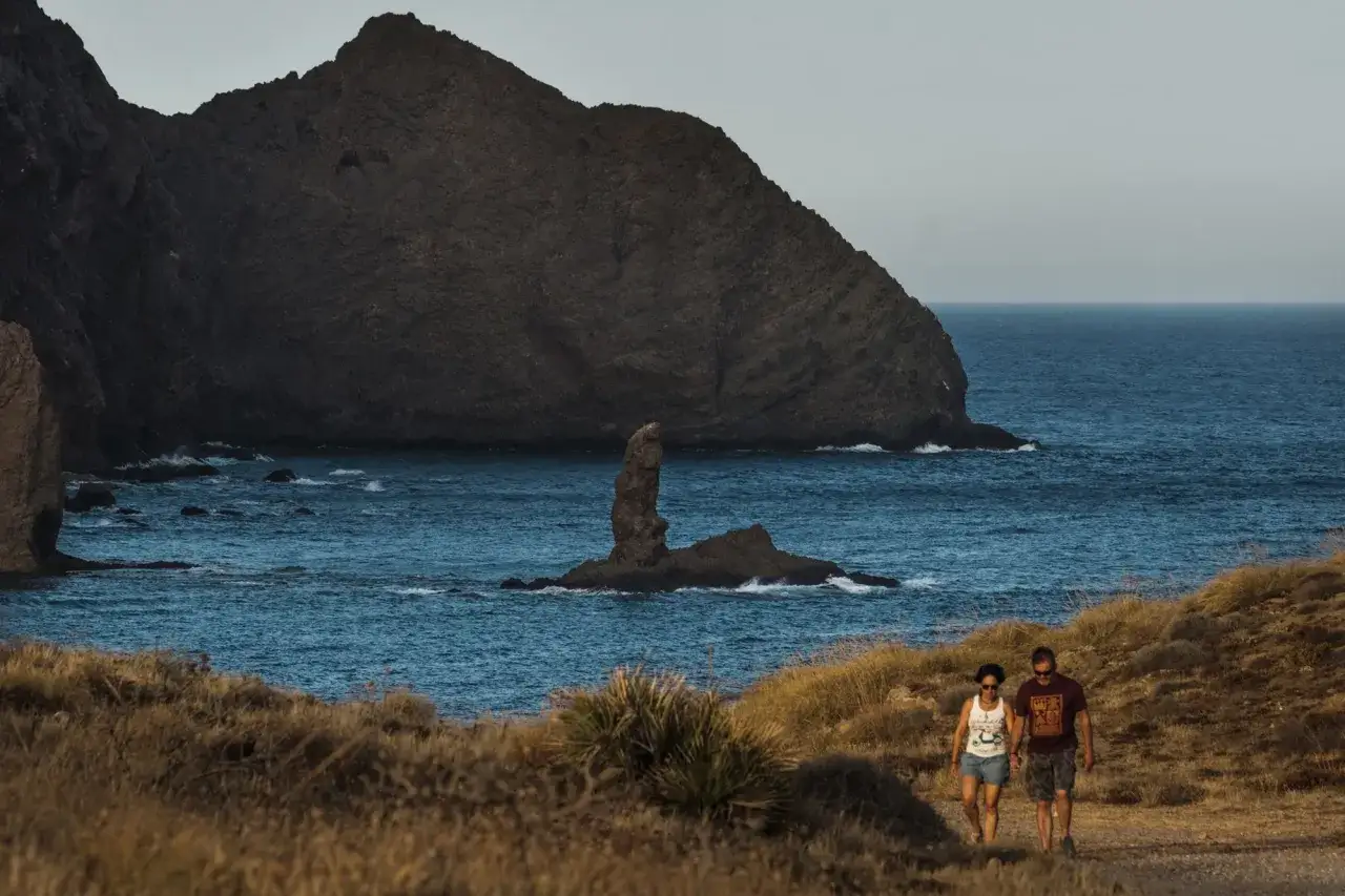 Pareja pasea cerca de la punta de loma pelada, con el mar azul y rocas escarpadas de fondo.