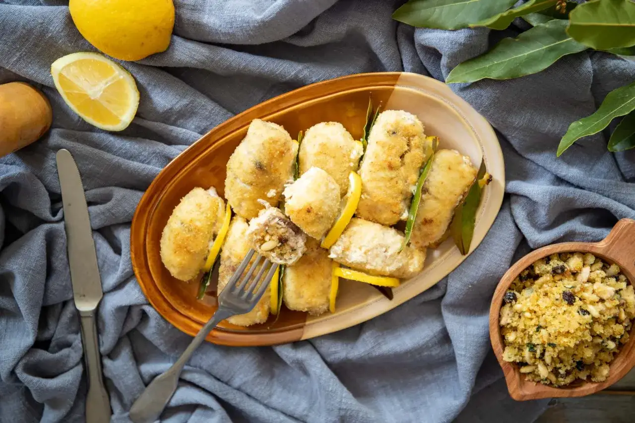 Deliziosi involtini di pesce spada impanati, guarniti con limone e alloro. Una forchetta solleva un involtino mostrando il ripieno saporito.