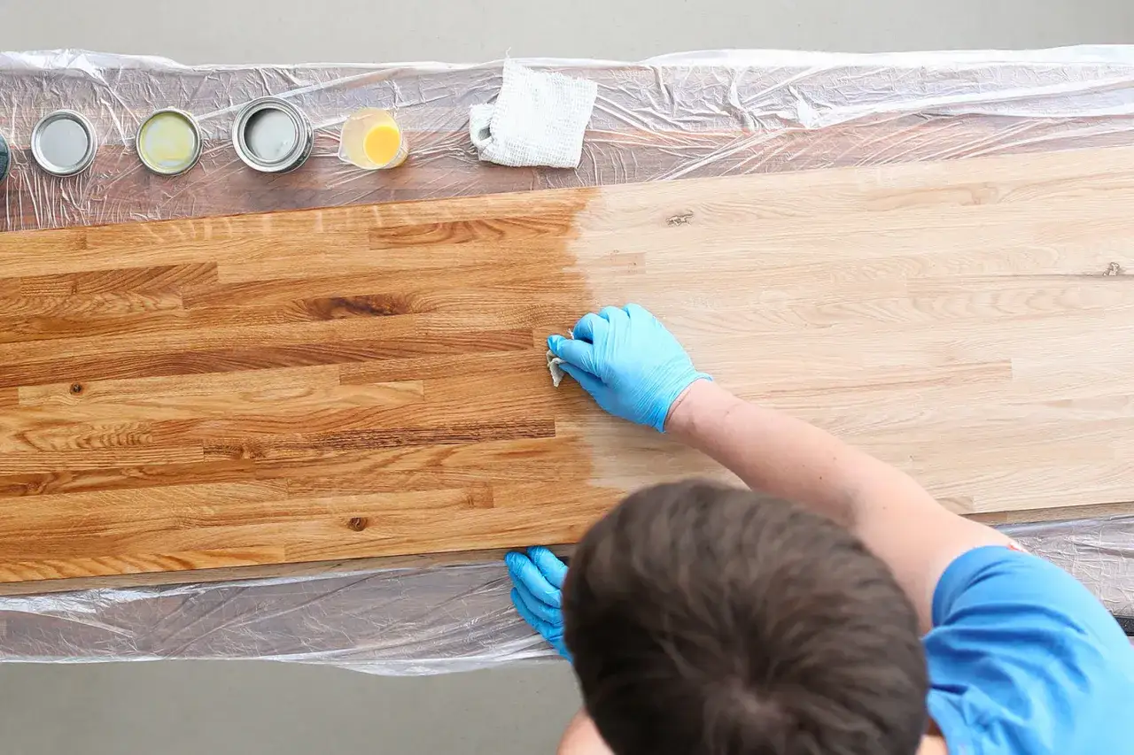 Mani con guanti blu applicano olio su un piano di legno, mostrando come smacchiare il legno e proteggerlo.