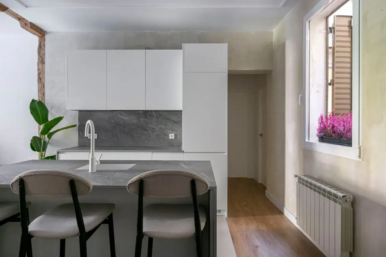 Cocina moderna con encimeras de cuarzo y **acabado fratasado**. Isla central con taburetes, grifer&iacute;a blanca y planta verde. Paredes texturizadas y ventana con flores.