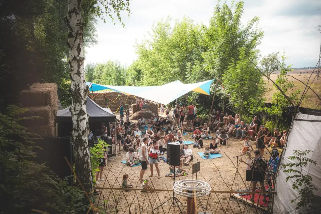 Menschen genie&szlig;en Musik und Gemeinschaft beim **Auerworld Festival** unter einem bunten Sonnensegel, umgeben von Strohballen und B&auml;umen.