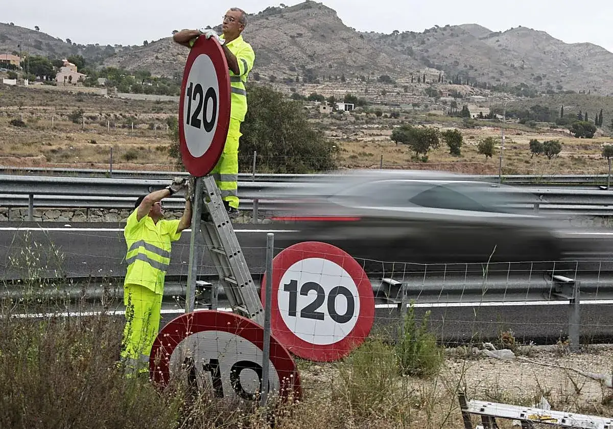 Dos operarios cambian señales de tráfico. Uno en escalera, ajusta un cartel de 120 km/h. Abajo, otro cartel marca 110. Evita la multa por ir a 140 en zona de 120.