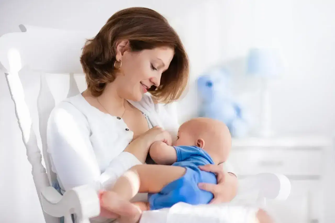 Uśmiechnięta mama karmi piersią dziecko w niebieskim ubranku. Mimo że maluch ma refluks u niemowlaka, czuje się bezpiecznie i kochany.