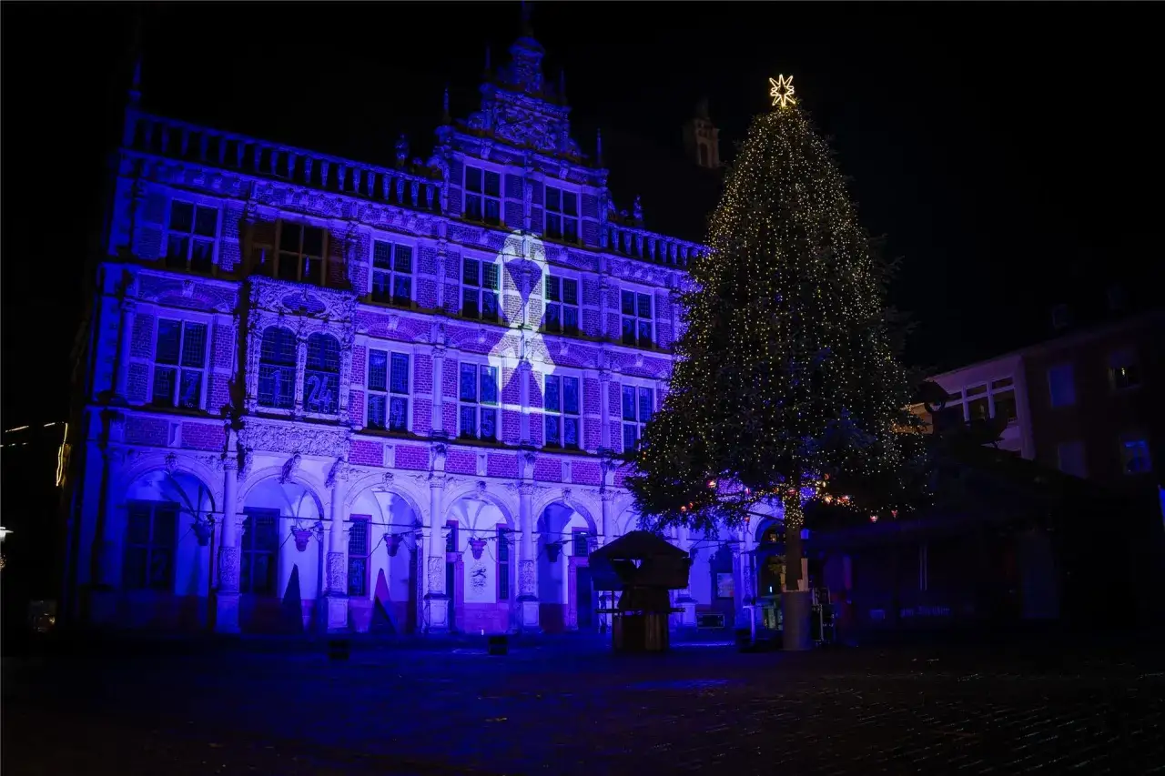 Festlicher Weihnachtsmarkt Bocholt: Ein pr&auml;chtiger, beleuchteter Weihnachtsbaum steht vor dem Rathaus, das in blaues Licht getaucht ist und eine wei&szlig;e Schleife projiziert.