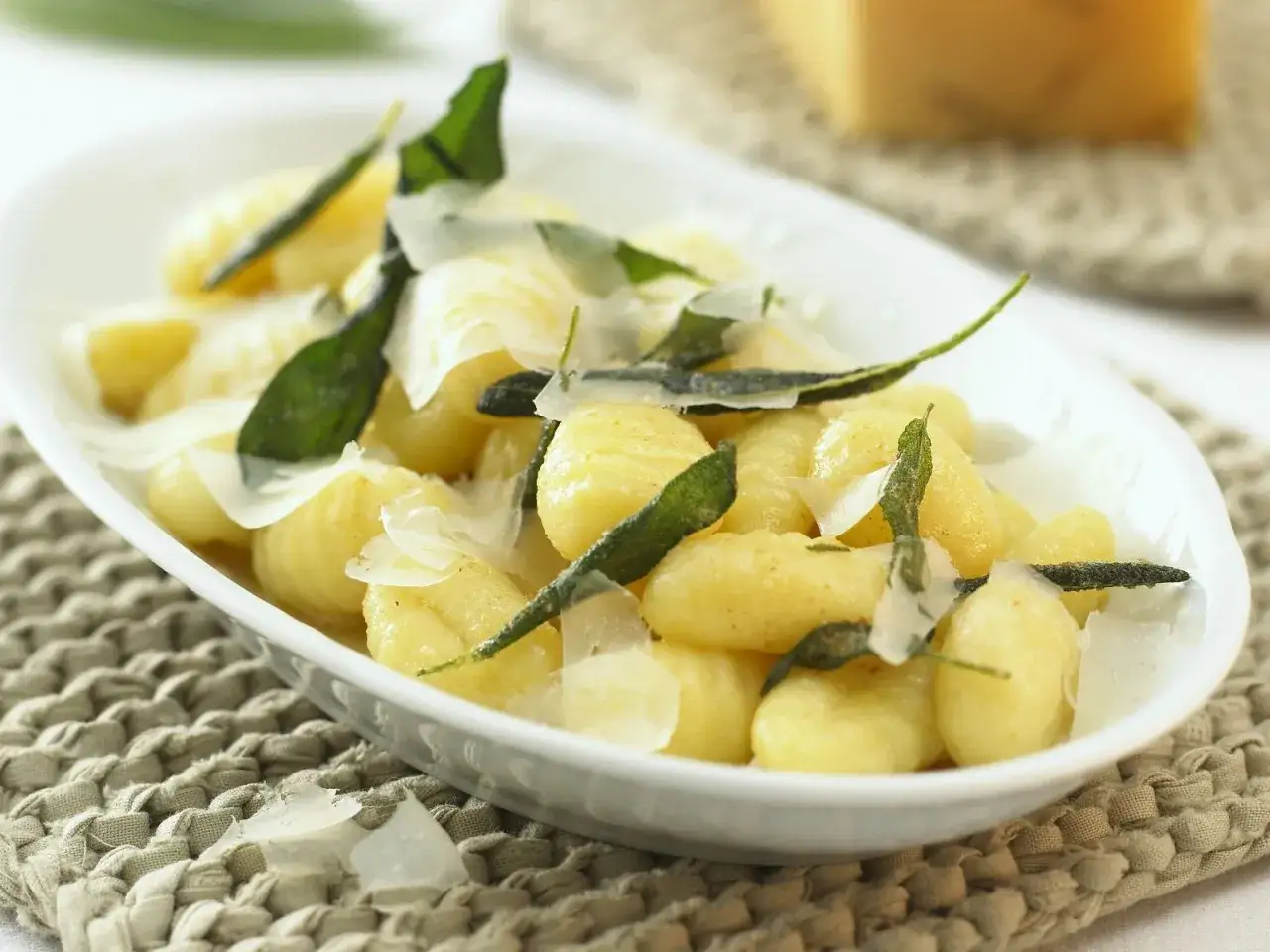 Salbei Gnocchi mit Parmesanflocken und frischen Salbeiblättern in einer weißen Schale auf einem geflochtenen Untersetzer.