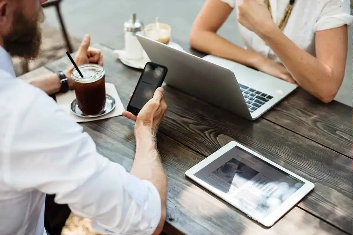 Dos personas trabajando en un caf&eacute;, con un port&aacute;til, tablet y m&oacute;vil. Un ejemplo de **trabajo n&oacute;mada digital**.
