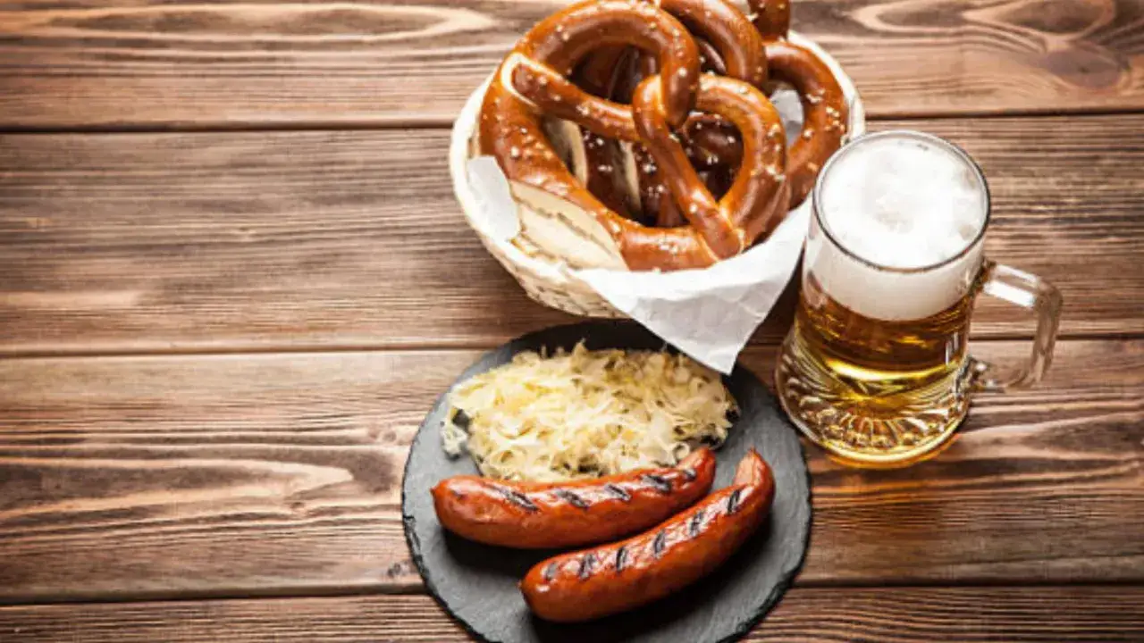 Un brindis por el oktoberfest valencia: salchichas a la parrilla, chucrut, pretzels y una jarra de cerveza espumosa sobre una mesa de madera.