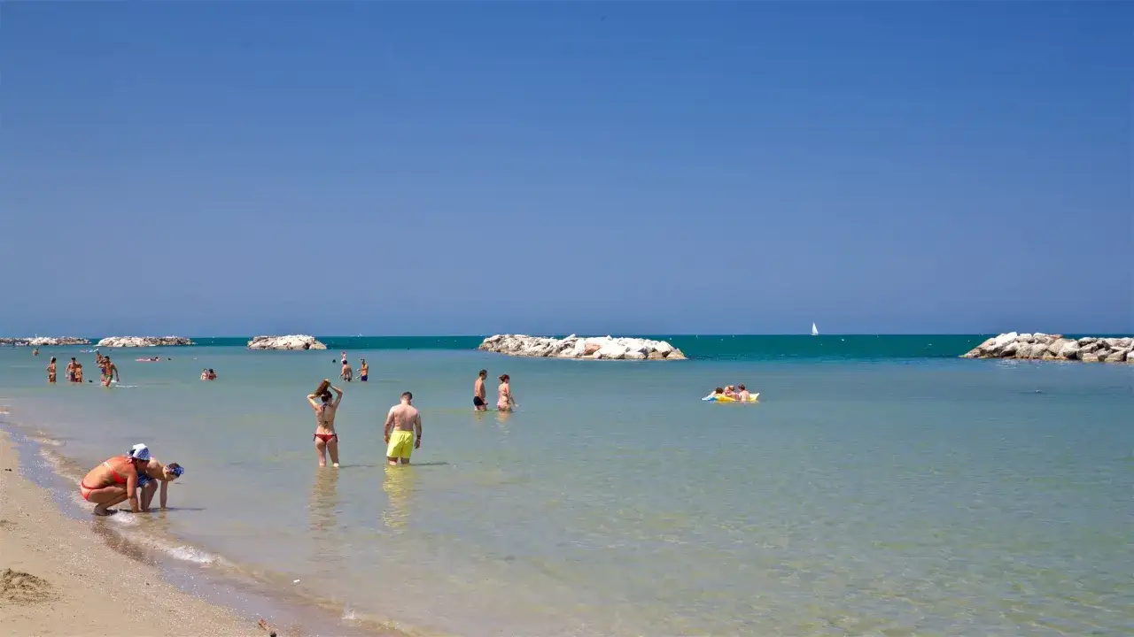 Spiaggia affollata con persone in acqua e sulla sabbia, vicino a una **torre pedrera** nel **centro storico**.
