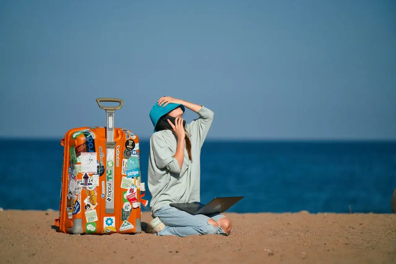 Frau sitzt am Strand mit Laptop und Koffer, telefoniert und wirkt gestresst. Sie ist krank im Urlaub und muss arbeiten.