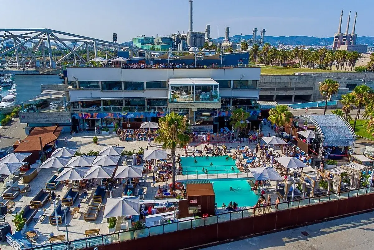 Un complesso di piscine e aree lounge con persone che si rilassano e si divertono, con un palco e uno sfondo industriale. Le discoteche Barceloneta offrono un'esperienza estiva vibrante.
