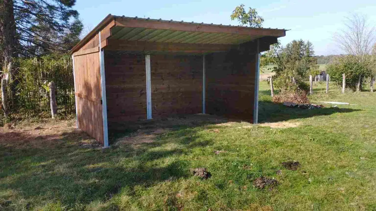 Abri en bois pour avoir un cheval chez soi, avec poteaux m&eacute;talliques et toit ondul&eacute;. L'herbe est verte autour.
