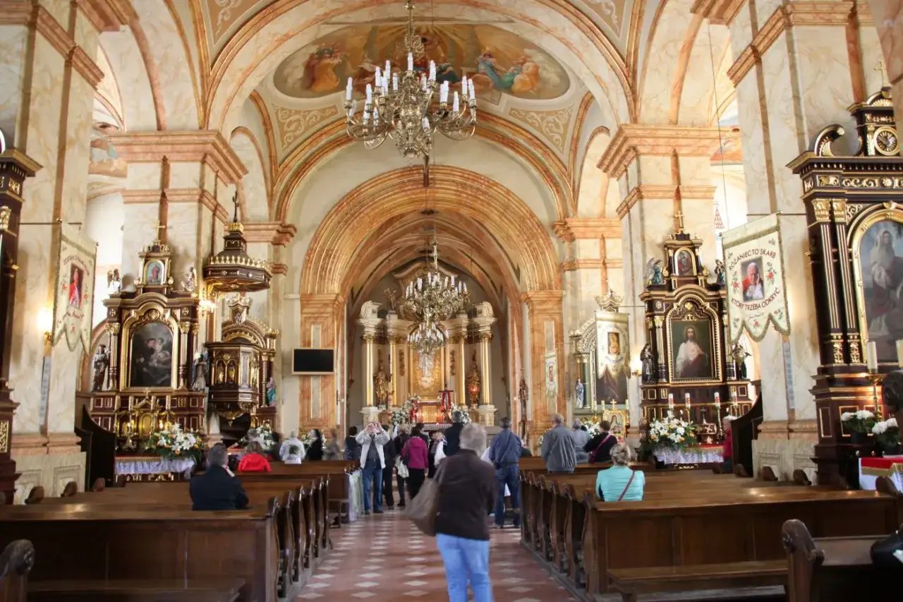 Wnętrze kościoła w Wadowicach, z wiernymi siedzącymi w ławkach i podziwiającymi bogate zdobienia, ołtarze i obrazy.