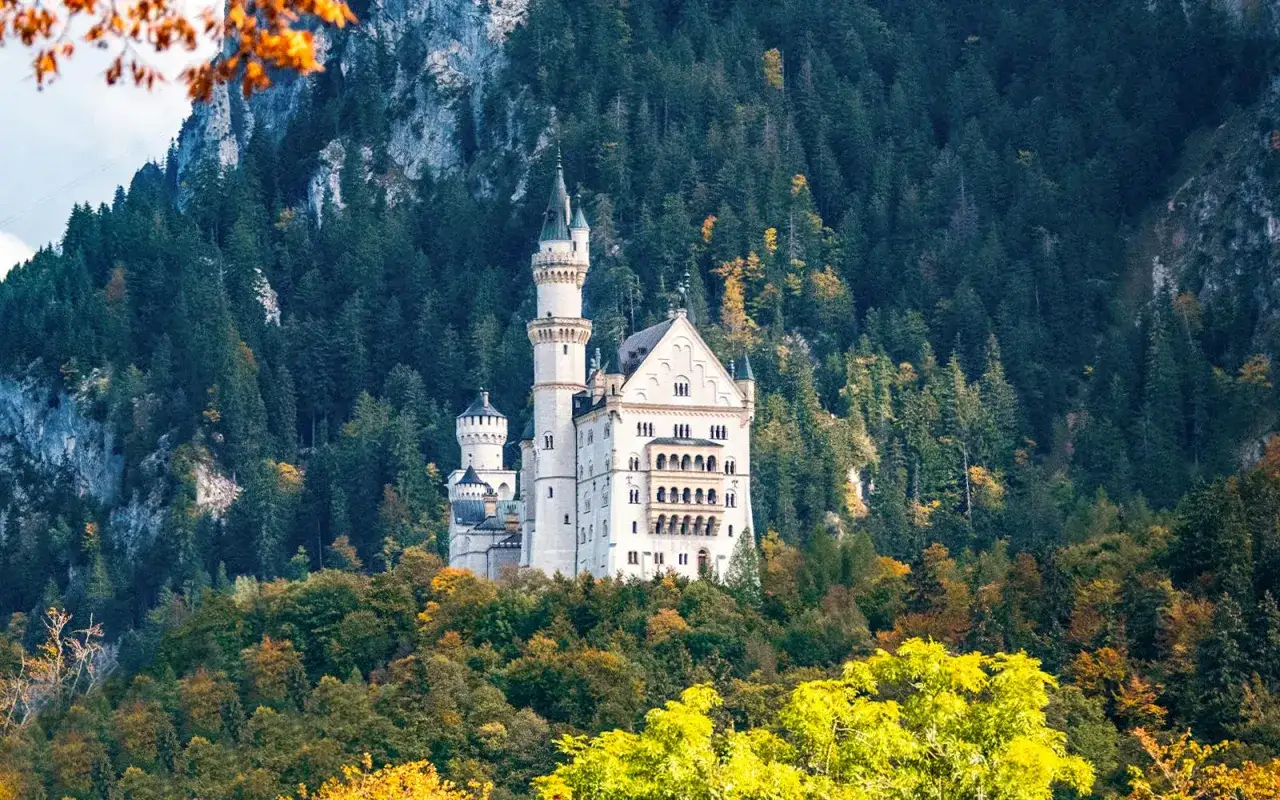 Neuschwanstein slott reser sig majestätiskt mot en bakgrund av tät skog och klippor. Höstfärgerna lyser upp träden runt det sagolika slottet.