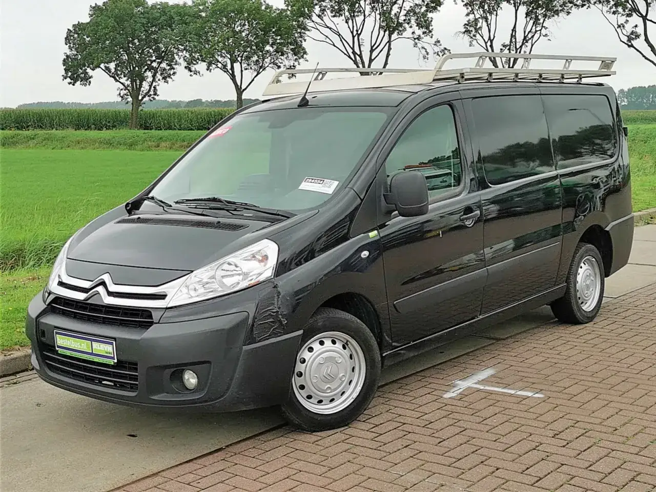 Czarny Citroen Jumpy 2.0 HDI z bagażnikiem dachowym, gotowy do pracy na tle zielonych pól i drzew.