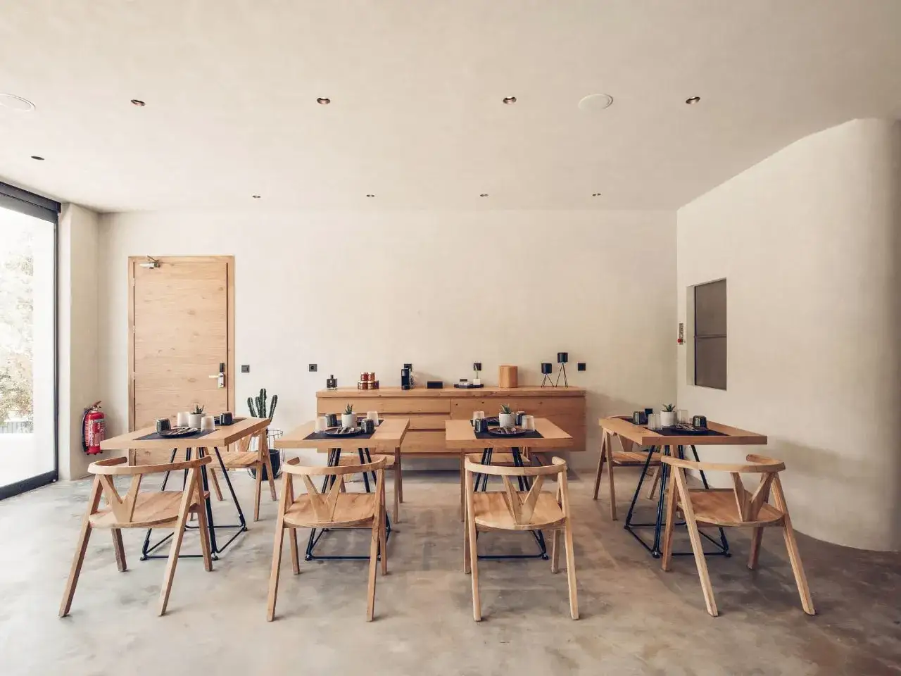 Minimalist dining area with wooden tables and chairs, ready for the reception and care of guests. A fire extinguisher stands by the door.