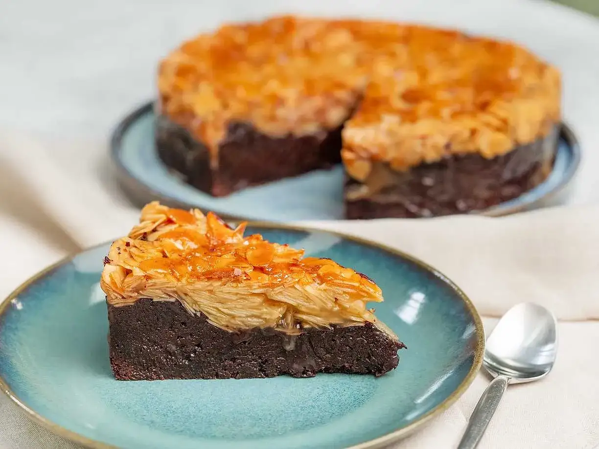 En bit chokladkaka med toscat&auml;cke av mandel. En hel toscakaka med chokladbotten syns i bakgrunden.