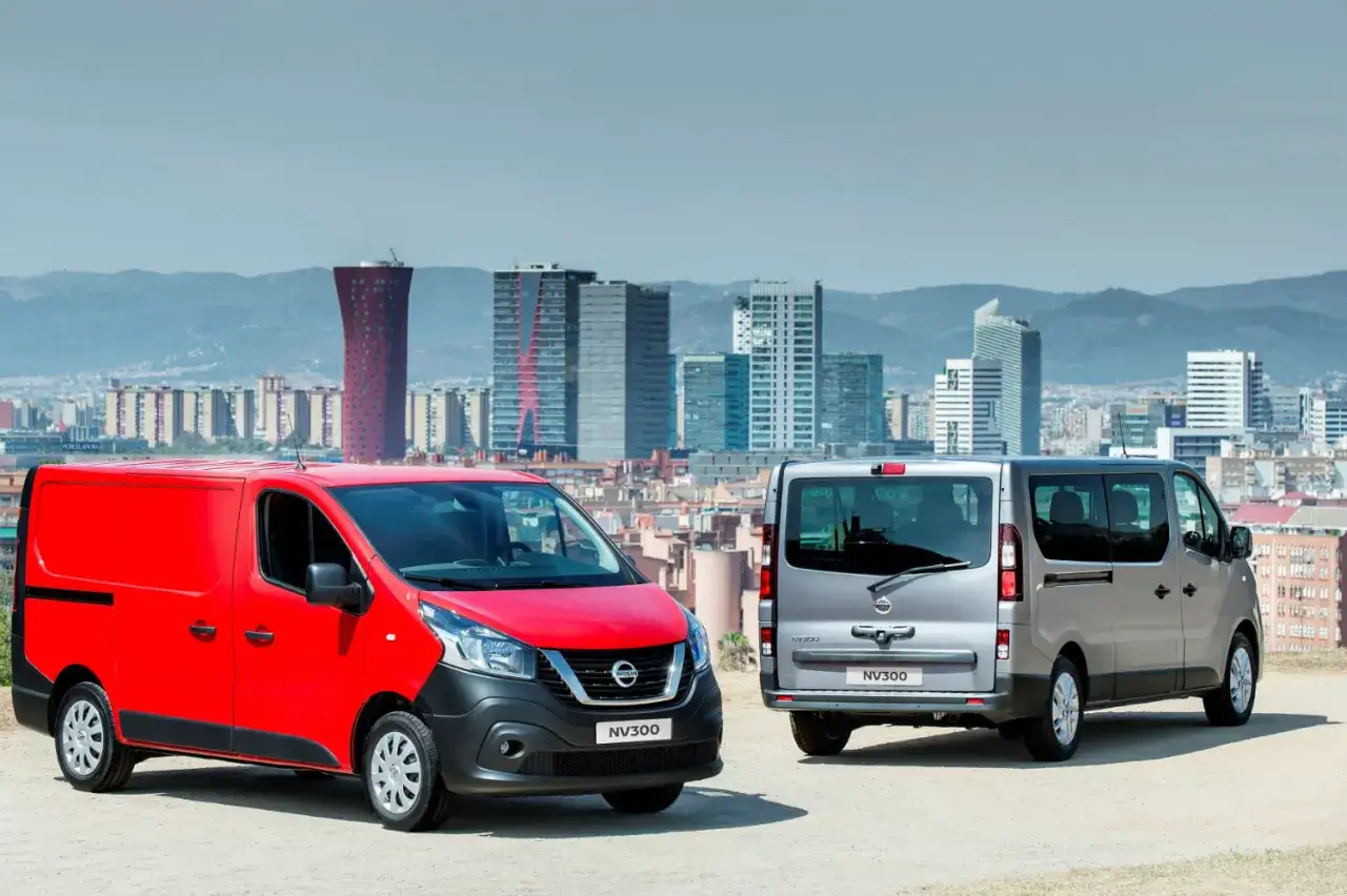 Dwa samochody dostawcze Nissan NV300: czerwony z przodu i srebrny z tyłu, na tle miejskiej panoramy z nowoczesnymi budynkami.