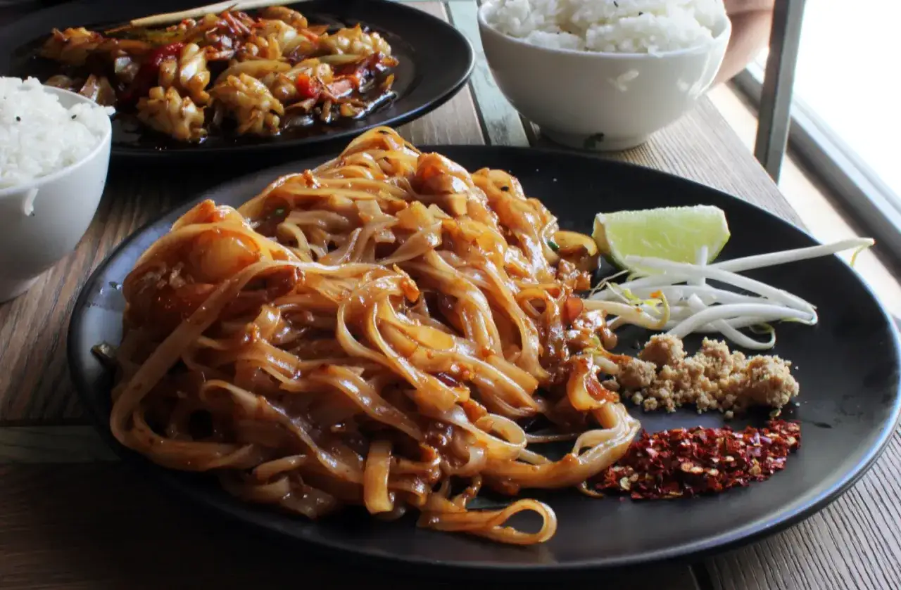 Talerz z makaronem pad thai, posypany orzeszkami ziemnymi i płatkami chili. Obok limonka i kiełki fasoli. Danie z tamaryndowca kusi smakiem.