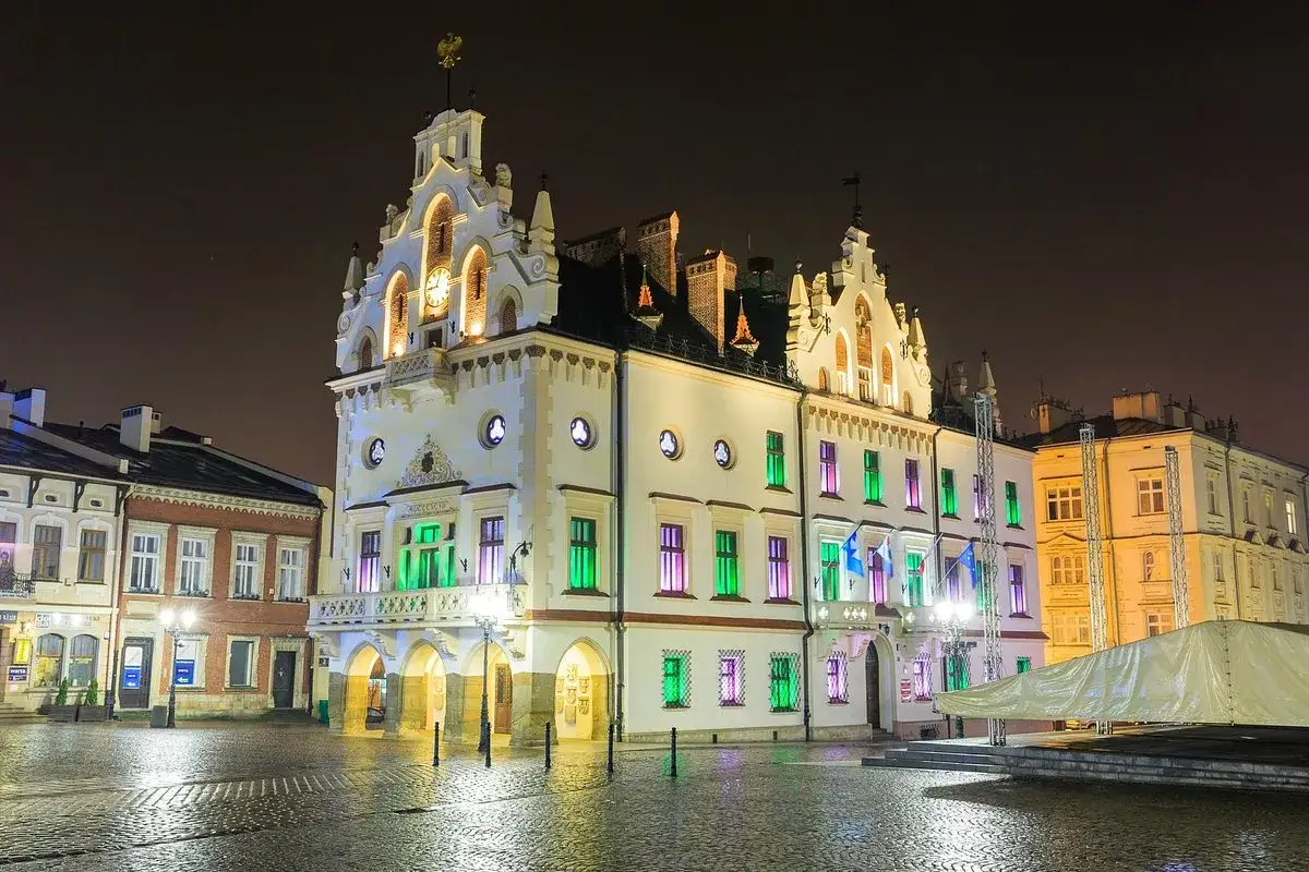 Rzeszów rynek nocą. Ratusz oświetlony kolorowo, z zegarem na wieży. Mokry bruk odbija światła.