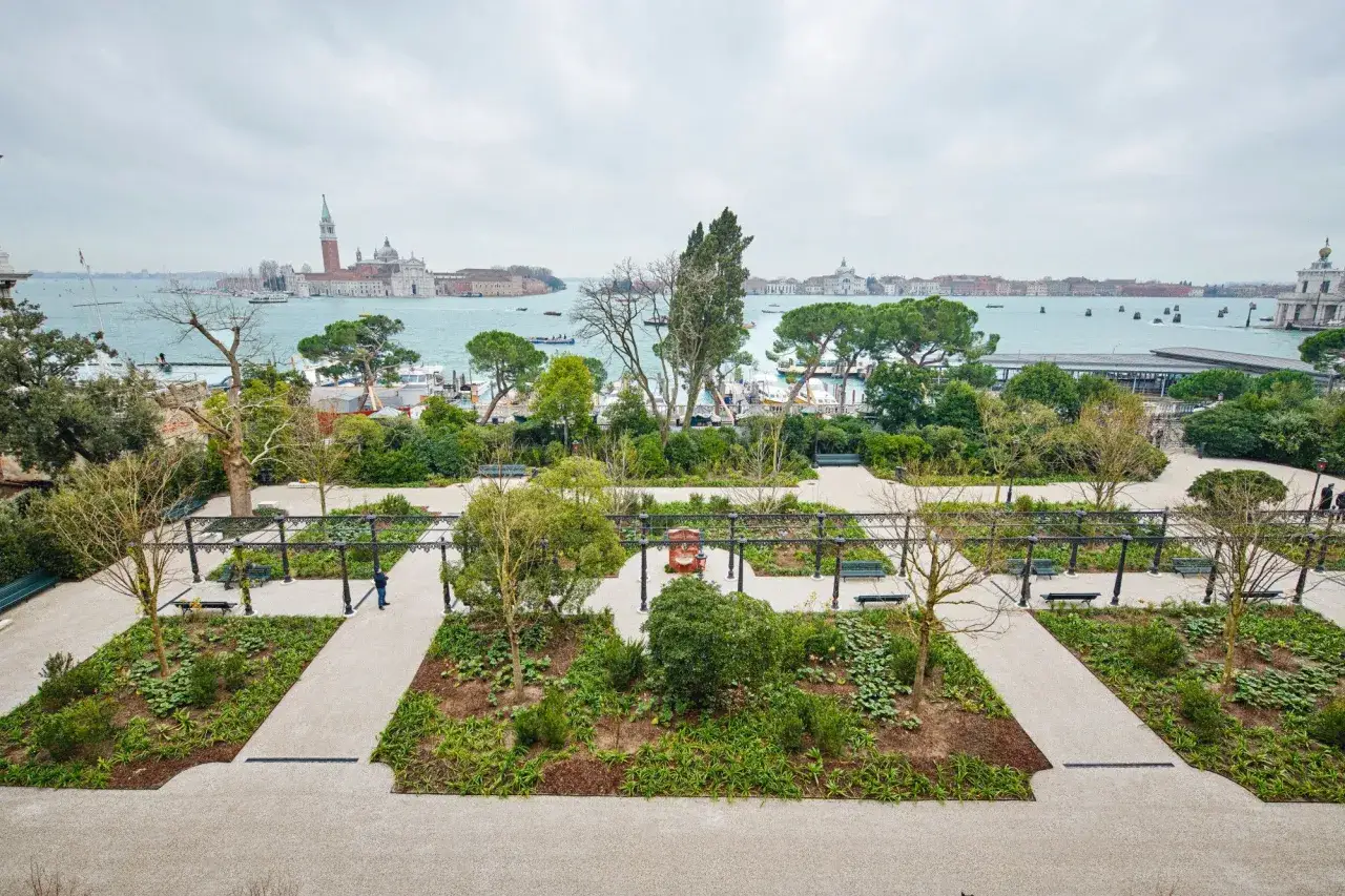 Vue d'un jardin &agrave; Venise, avec des parterres de fleurs, des pergolas et des bancs, donnant sur le lagon et les b&acirc;timents historiques.
