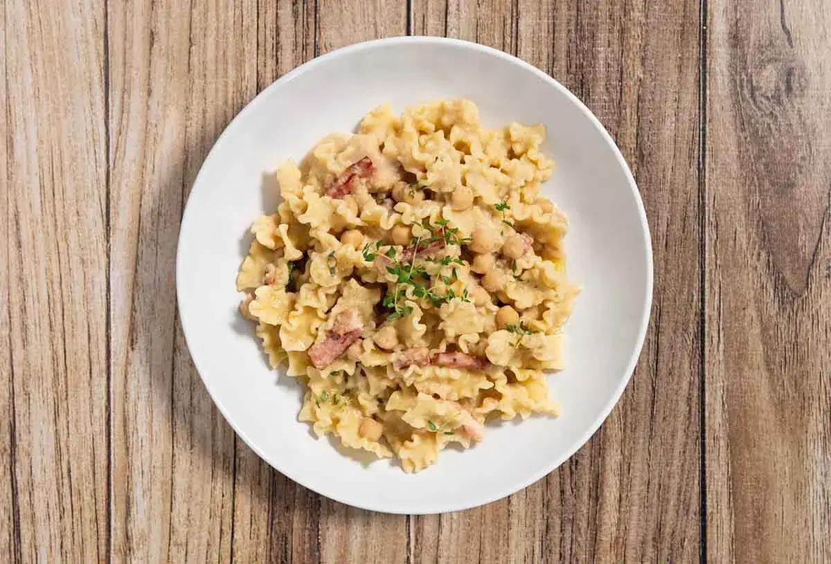 Un plato de pasta e ceci con garbanzos, trozos de panceta y hierbas frescas, servido en un cuenco blanco sobre una mesa de madera.