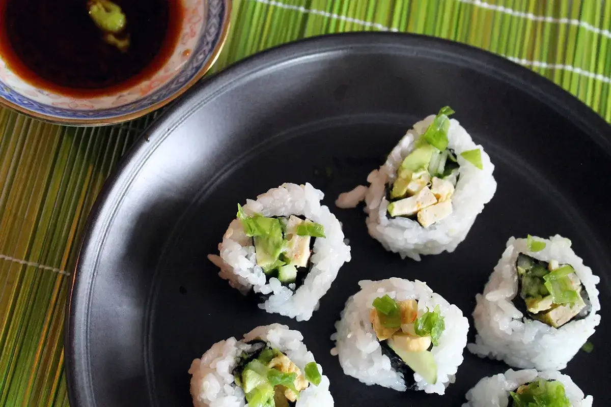 Kilka kawałków california sushi z awokado i ogórkiem na czarnym talerzu, obok miseczka sosu sojowego z wasabi.