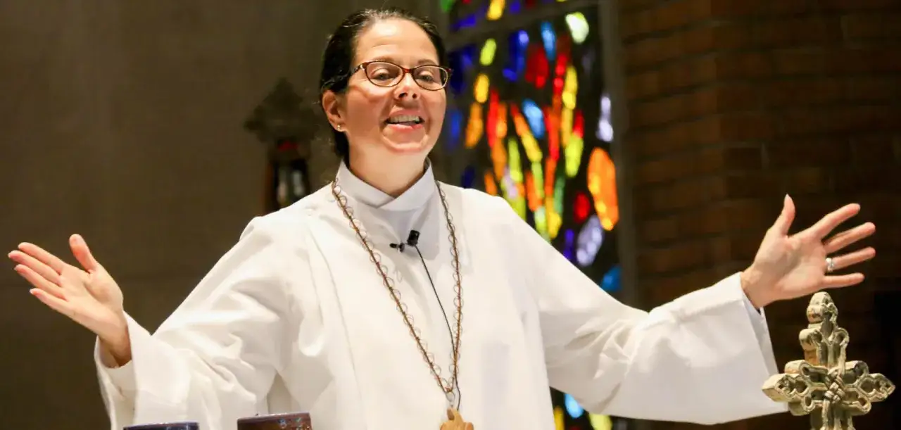 A woman in a white robe, wearing glasses and a cross necklace, speaks with open hands, demonstrating that women can be pastors.