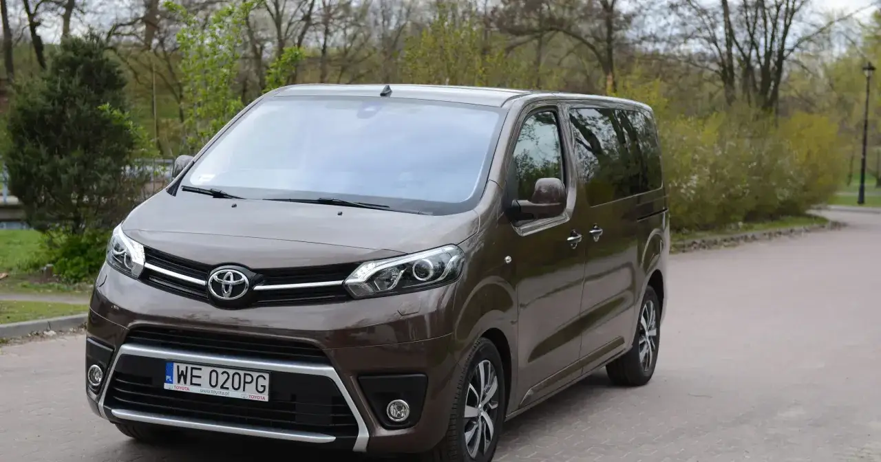Brązowy Toyota van zaparkowany na wybrukowanym podjeździe, otoczony zielenią.