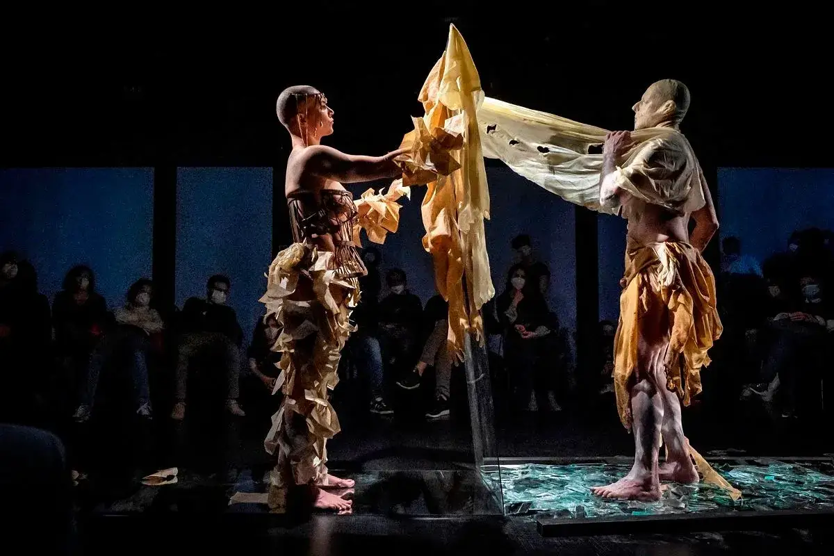 Dwie postacie w teatralnych strojach z papieru i tkaniny, tworzące scenę polskiej sztuki współczesnej. Widownia w maskach obserwuje performance.