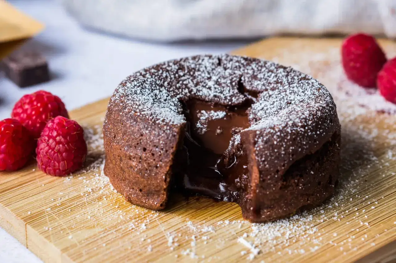 Un fondant au chocolat coulant, saupoudré de sucre glace, avec des framboises fraîches sur une planche en bois.