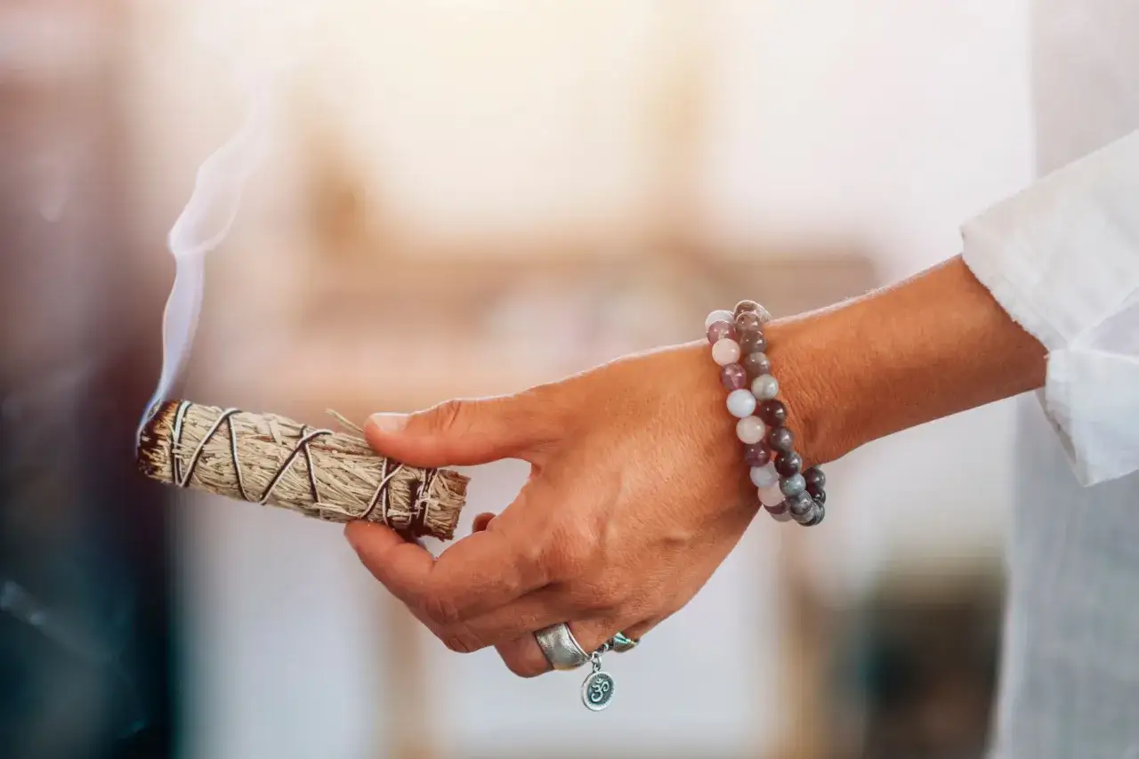Mano con pulsera de piedras sostiene un manojo de salvia humeante, listo para sahumar la casa y purificar el ambiente.