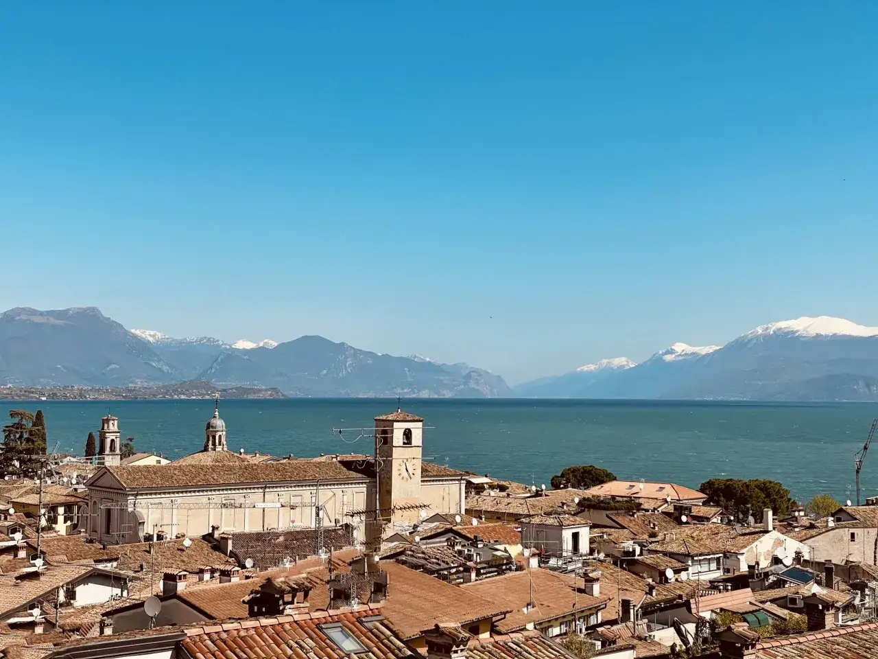 Tetti rossi e campanili nel centro storico di Desenzano del Garda, con il lago e le montagne innevate sullo sfondo.