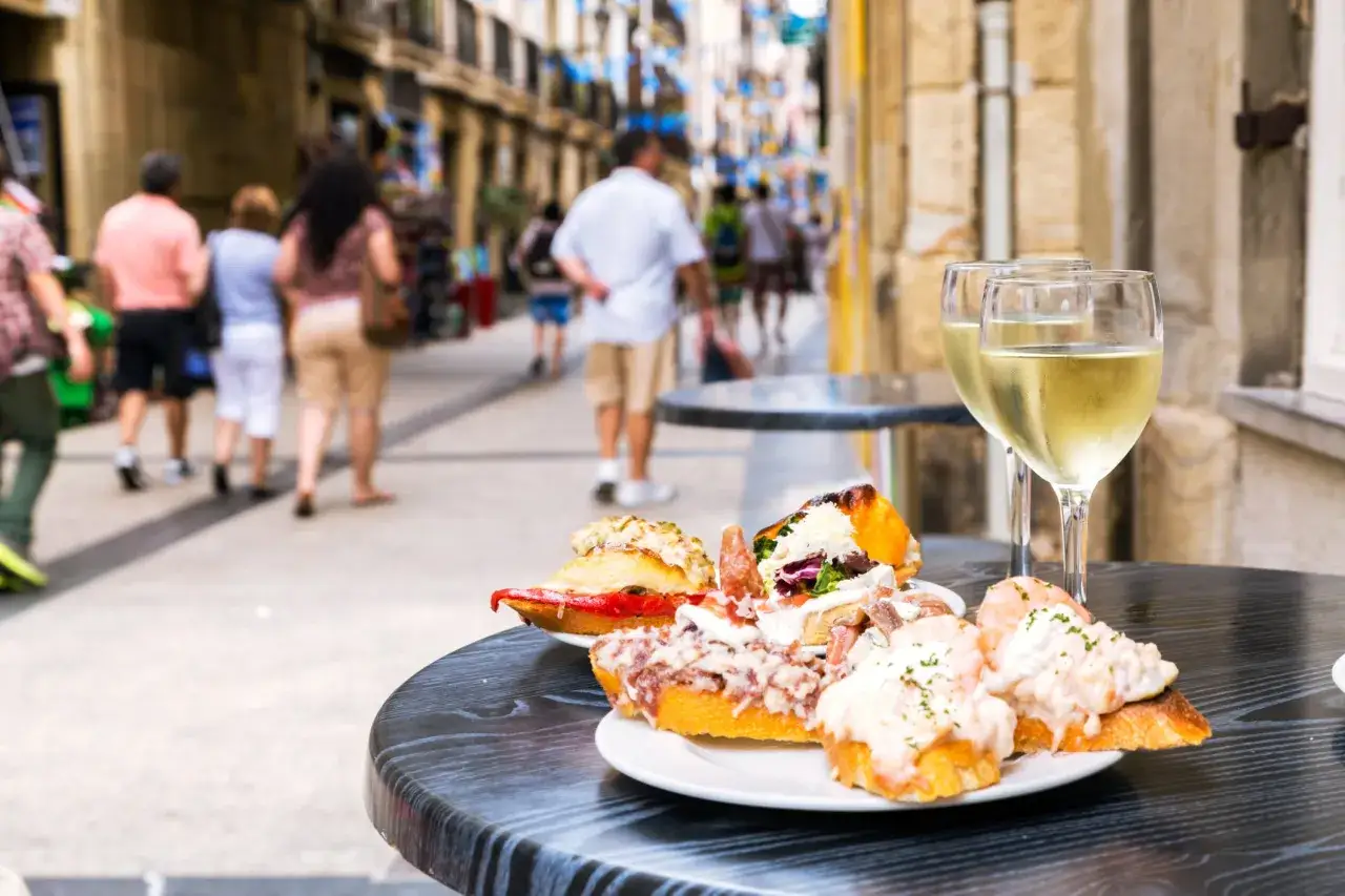 Tv&aring; glas vitt vin och en tallrik med l&auml;ckra pintxos p&aring; ett bord utanf&ouml;r en restaurang i San Sebasti&aacute;n. Folk promenerar f&ouml;rbi p&aring; den livliga gatan.