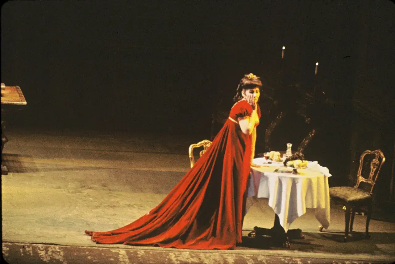 Maria Callas w czerwonej sukni, przy stole z owocami, scena z opery. Jej biografia pełna jest takich dramatycznych momentów.