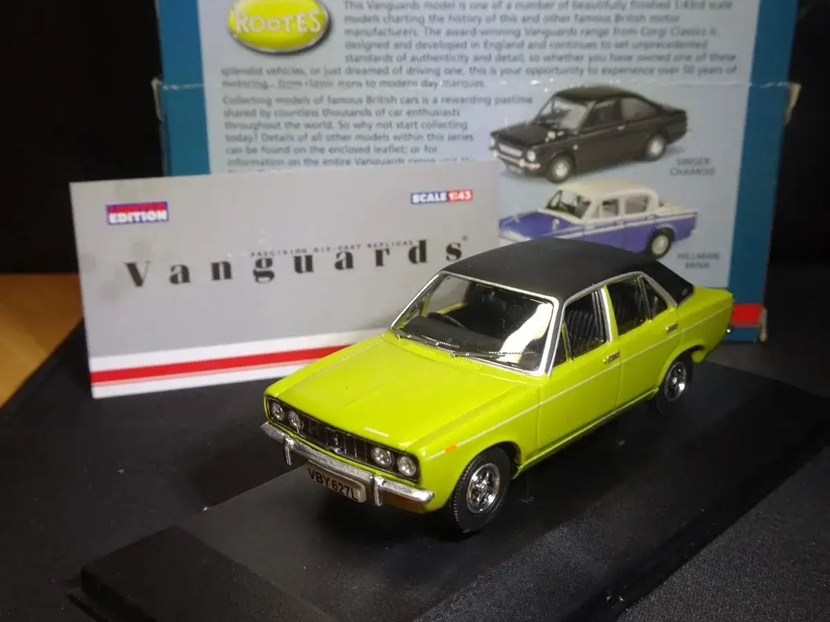 En gulgr&ouml;n Hillman Minx-modellbil st&aring;r p&aring; ett svart stativ. I bakgrunden syns en kartong med text om Vanguards-modeller.