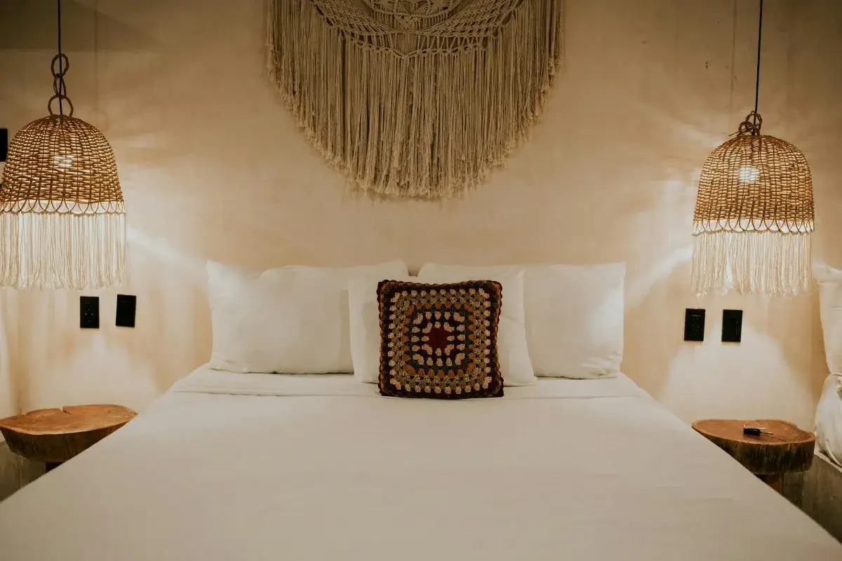 Un dormitorio eclectico con cama blanca, cojín de ganchillo, lámparas de mimbre y macramé en la pared.