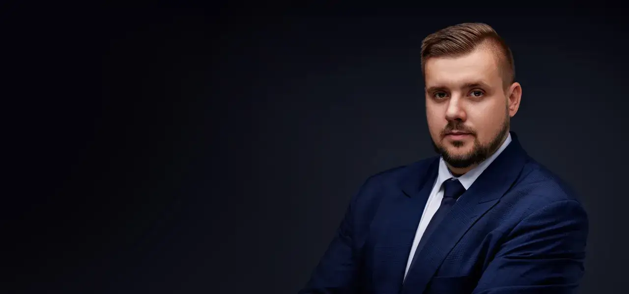 Portret mężczyzny w granatowym garniturze i krawacie, na ciemnym tle.