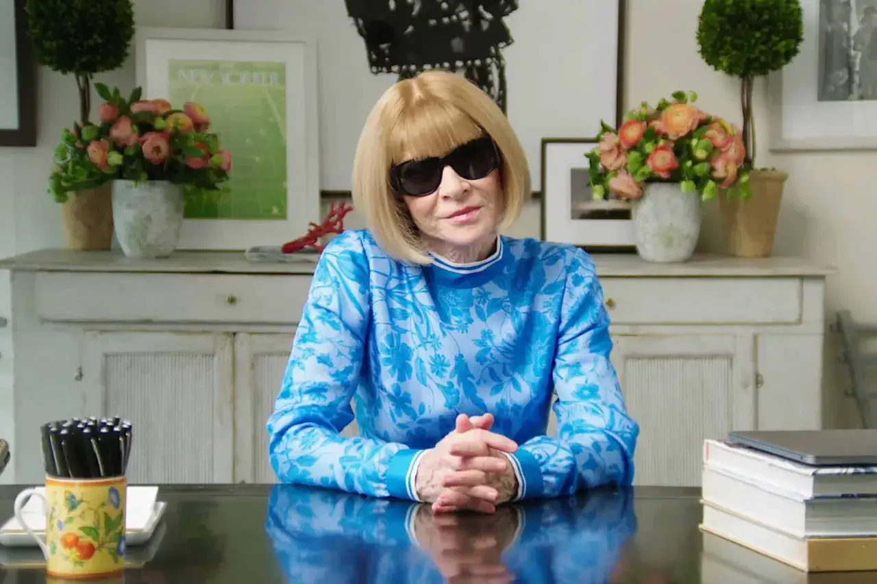 Anna Wintour, ikona Vogue, w niebieskiej bluzce z kwiatowym wzorem, siedzi przy biurku.