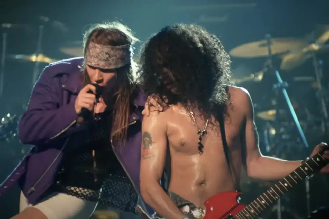 Axl Rose z zespołem Guns N' Roses na scenie. Wokalista w fioletowej kurtce śpiewa, a Slash gra na gitarze.