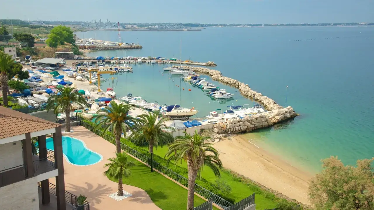 En vacker vy över en hamn med många båtar i Taranto, Italien. En pool och palmer syns i förgrunden.