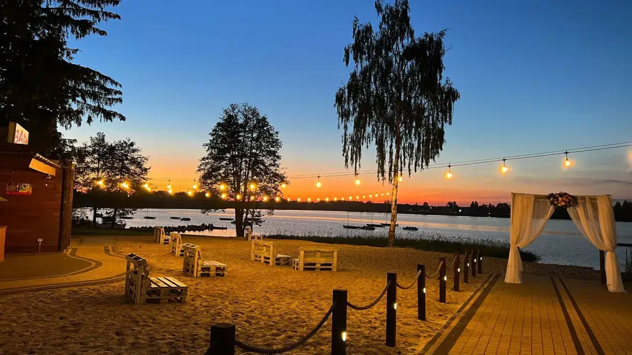 Piękny zachód słońca nad jeziorem. Plaża z drewnianymi meblami i girlandą świateł, idealne miejsce na relaks. Okuninka atrakcje dla dzieci czekają!
