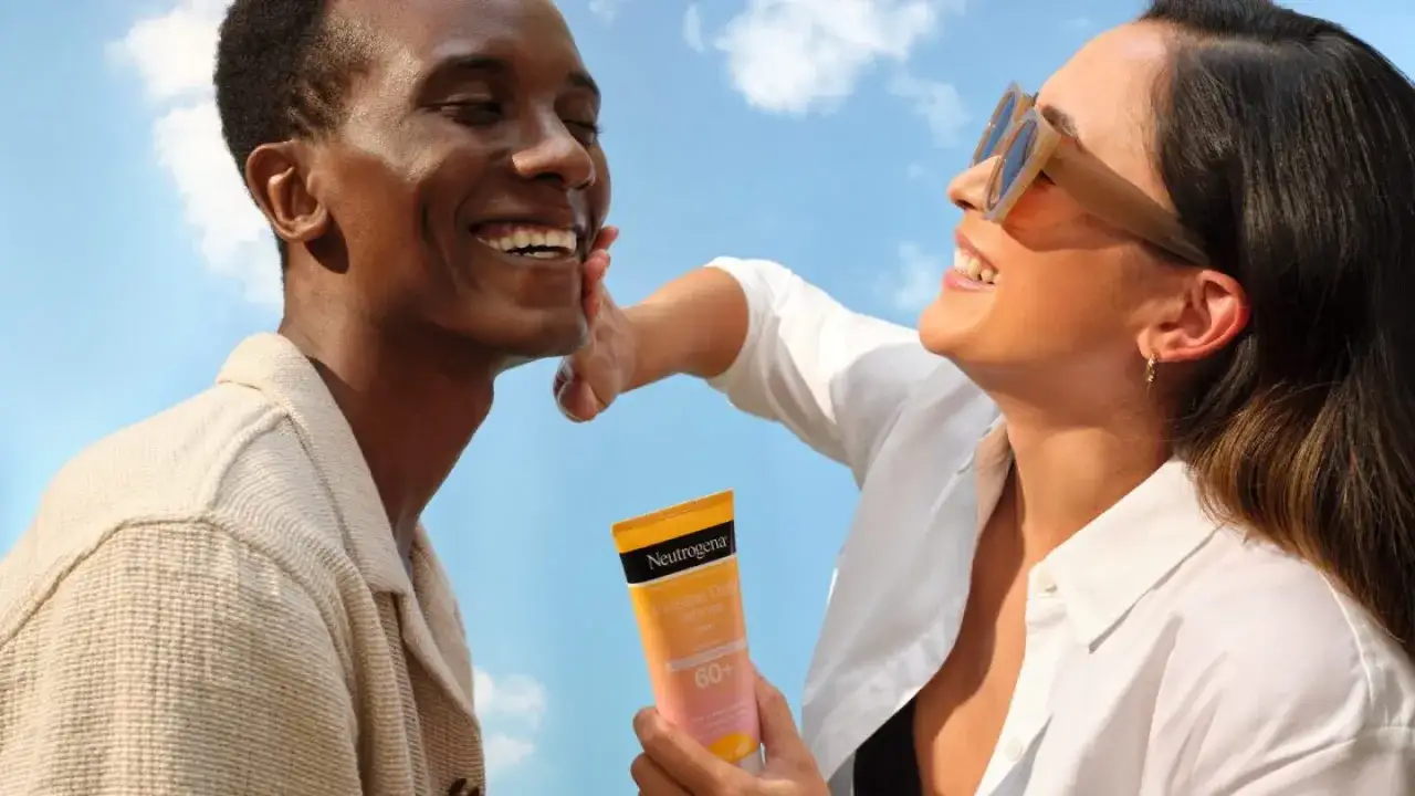 Pareja sonriente bajo el sol, ella aplica protector solar con factor de protección solar 60+ en el rostro de él.