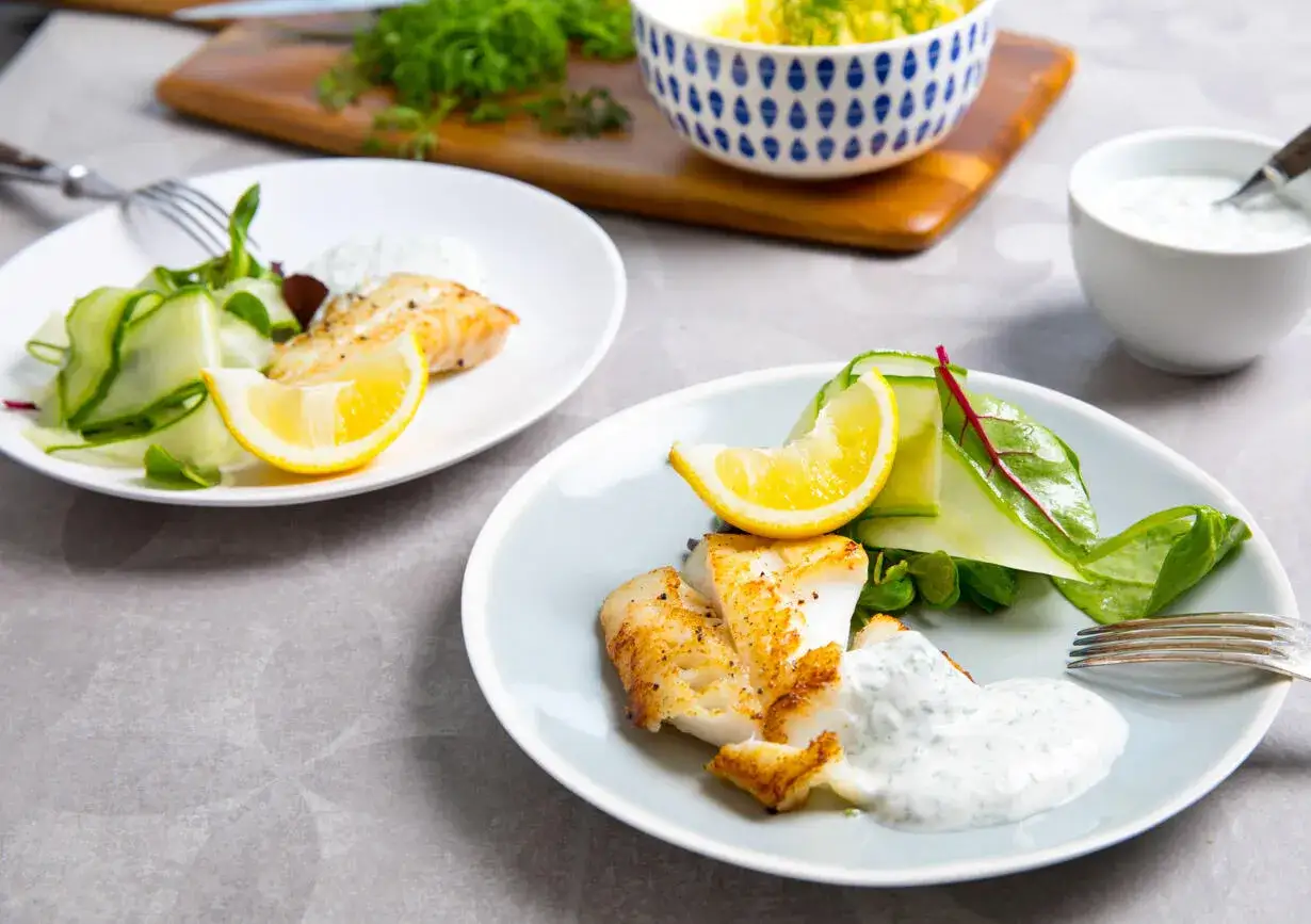 Två tallrikar med stekt torskfile, serverad med citronskiva, gurksallad och en krämig sås.