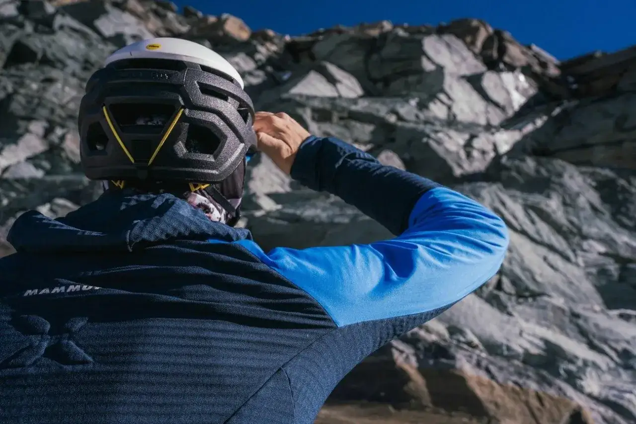 A hiker in a helmet and blue jacket surveys the rugged terrain, ready for alpine hiking.