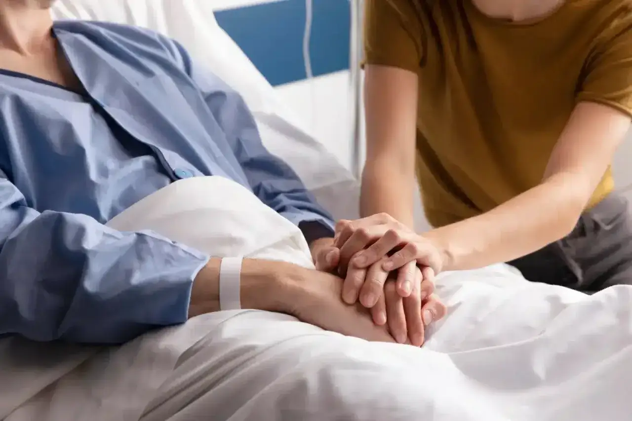 Osoba w szpitalnej piżamie trzyma dłoń bliskiej osoby. To obraz wsparcia i troski w trudnych chwilach, gdzie leczenie paliatywne zapewnia komfort.