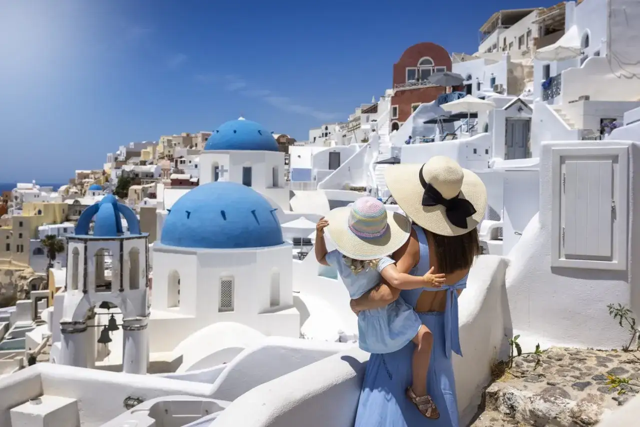 Una madre abbraccia la sua bambina, ammirando il panorama di Santorini, Grecia. Le cupole blu brillano sotto il sole, un ricordo indimenticabile di vacanza con bambini.