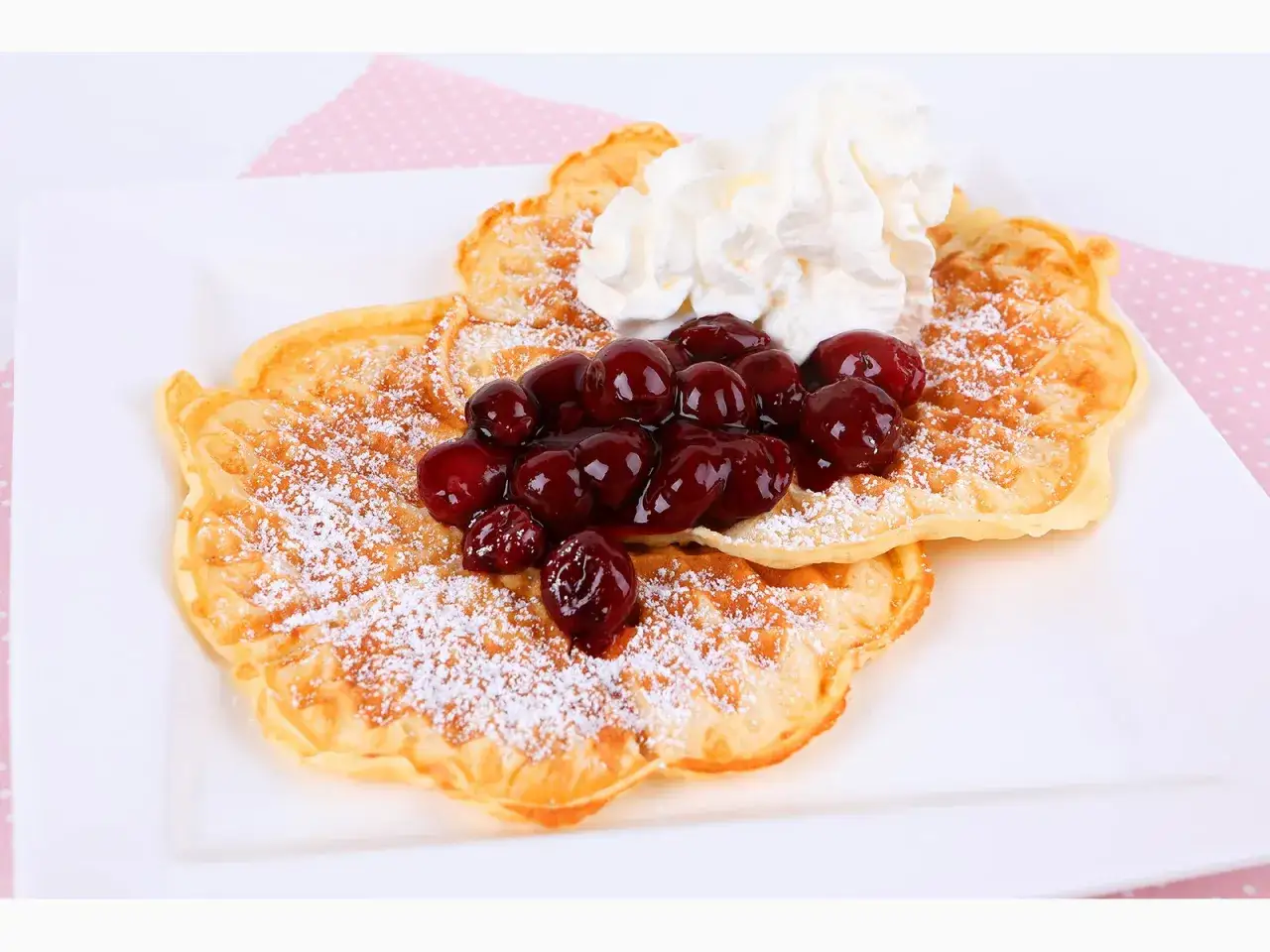Zwei goldbraune Waffeln, bestäubt mit Puderzucker, belegt mit Kirschen und Sahne. Ein köstliches Waffeln Rezept für den Genuss.