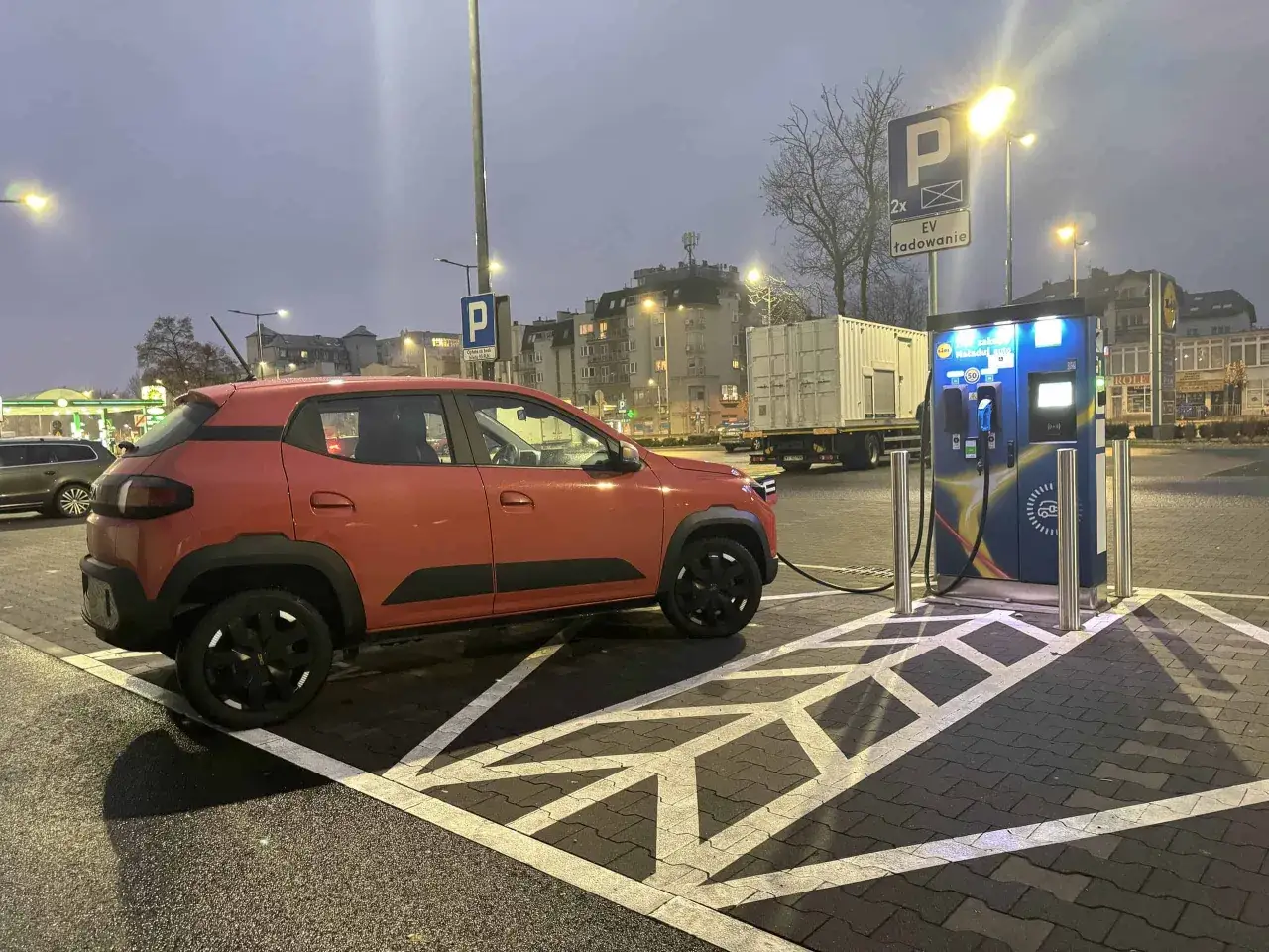 Pomarańczowy Dacia Spring ładuje się na stacji EV. Znak 